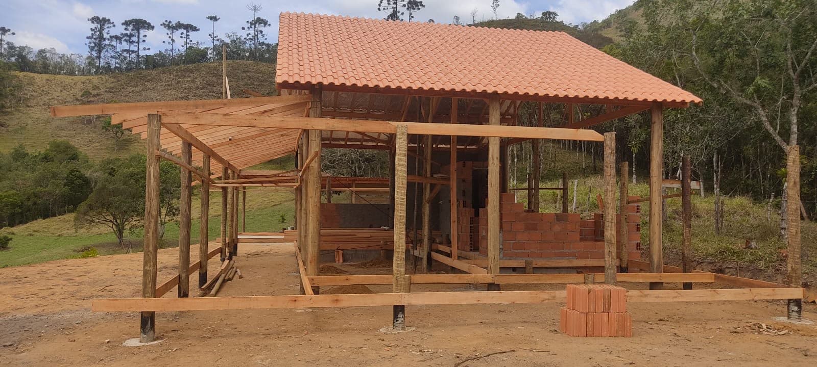 Obra em estrutura de madeira roliça com telhado cerâmico recém-assentado, entre morros da serra